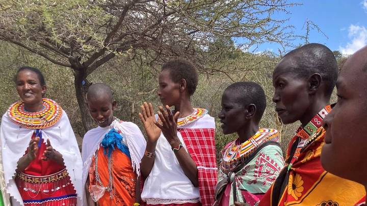 A row of Maasai women in vibrant traditional beadwork and brightly colored shukas clapping and singing outdoors under acacia trees.
