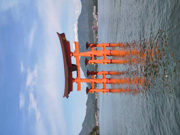 Iconic bright vermilion torii gate of Itsukushima Shrine standing in calm blue sea water with distant mountains and town on the shore.