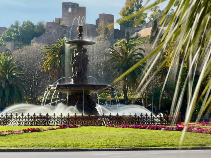Elegant city fountain surrounded by palms and flowerbeds with historic walls behind