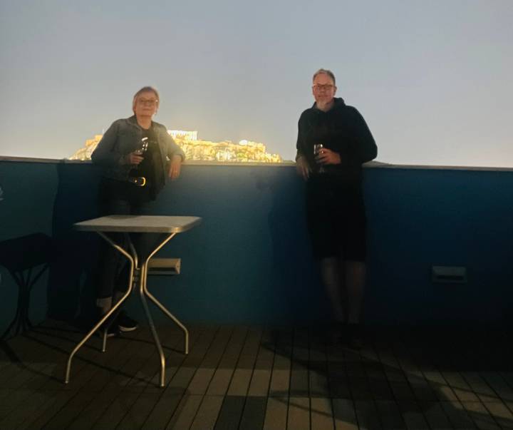 Very blurry nighttime roof-top photo of two people holding drinks with illuminated Acropolis in background