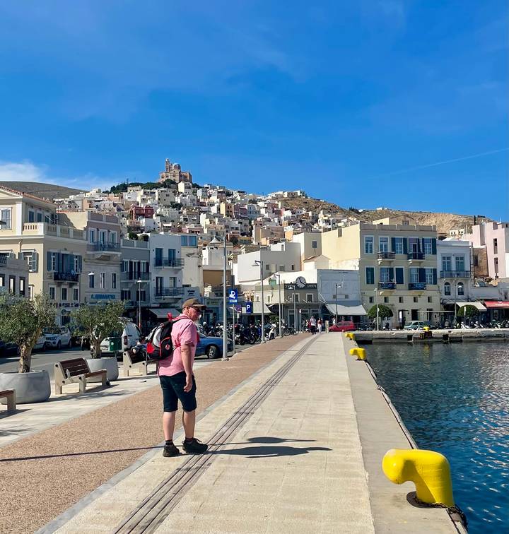 Promenade of Ermoupoli, Syros with pastel buildings on hillside and traveler walking waterside path