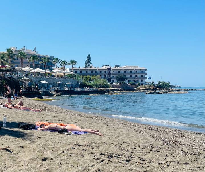 People sunbathing on a narrow sandy beach with calm Aegean waters and restaurants behind