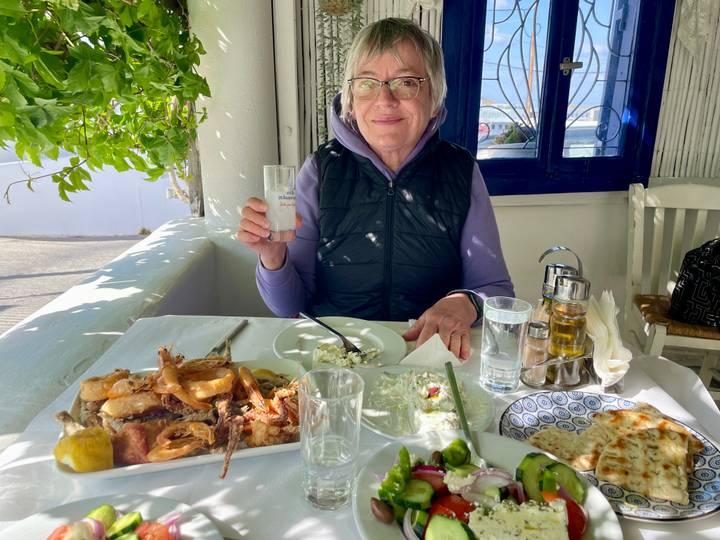 Smiling older woman enjoying a seafood feast at an outdoor Greek taverna with white walls and a blue-framed window.