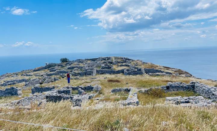 Stone ruins of an ancient hilltop settlement overlooking the deep-blue Aegean Sea with a single visitor wandering among the walls.