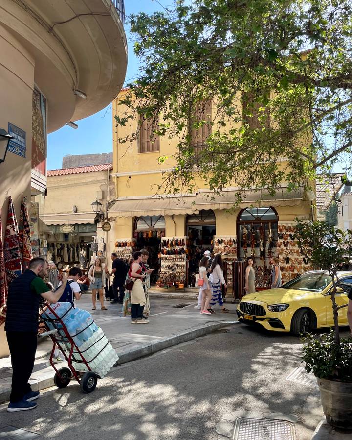 Bustling pedestrian street lined with leather-goods shops and café tables in a historic Greek town.