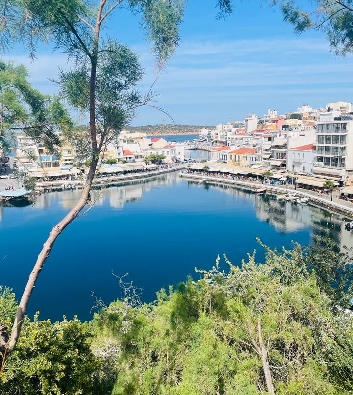 Bright view over a calm blue lake-style harbor ringed by whitewashed buildings with red roofs on Crete.