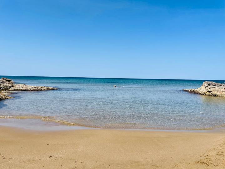 Quiet sandy cove with gentle turquoise water framed by rocky outcrops under a clear blue sky.