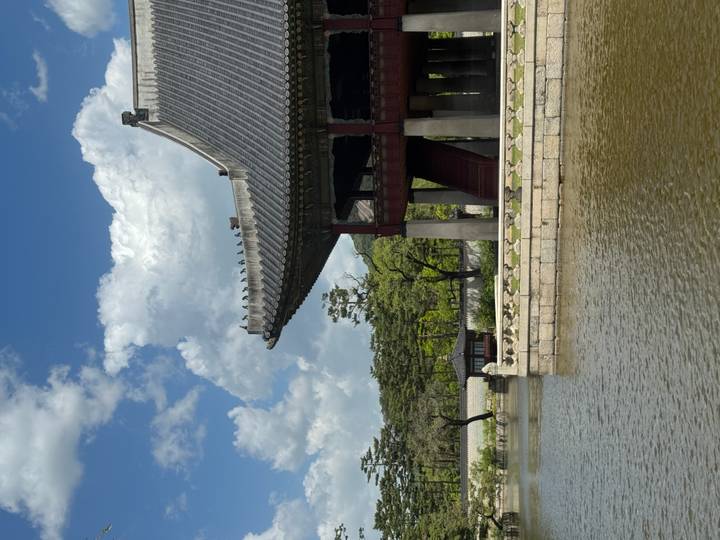 Traditional Gyeonghoeru pavilion over mirrored pond with forest backdrop and fluffy clouds