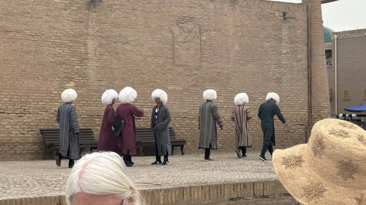 Men in traditional striped robes and large wool hats walking along mud-brick wall in Khiva