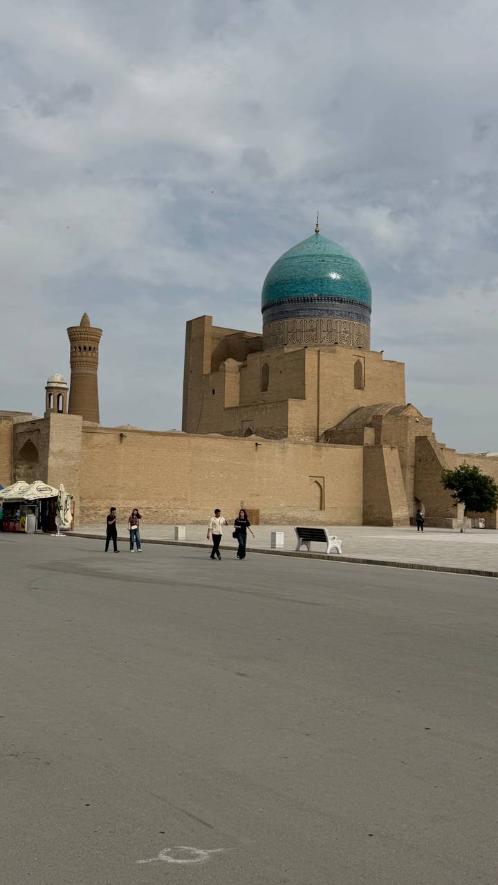 Historic brick minaret and mosque complex with a few visitors walking across a wide stone courtyard.