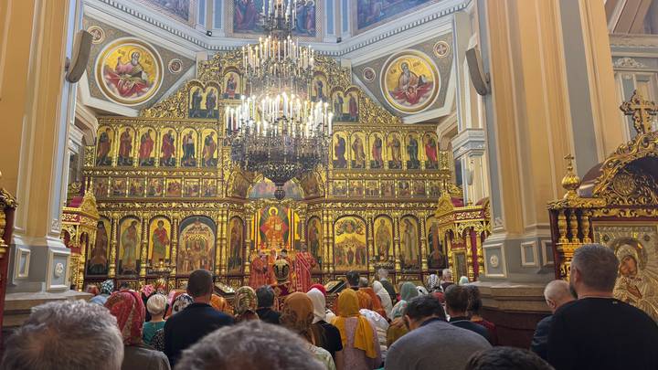 Lavish icon-covered interior of an Orthodox church packed with worshippers under a glittering chandelier.