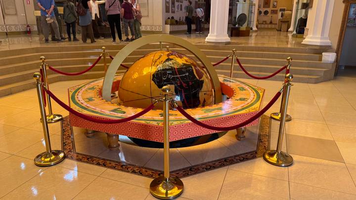 Large decorated globe display inside a museum rotunda cordoned by red ropes while visitors look on.
