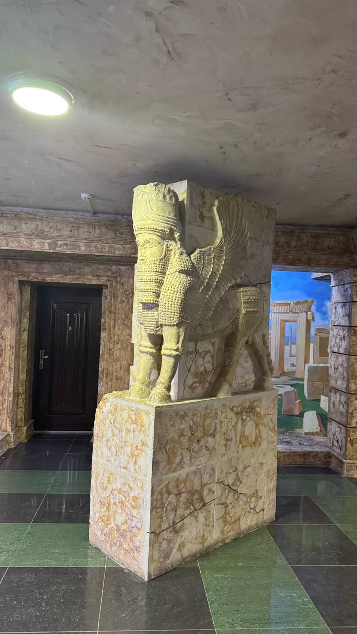 Stone Lamassu winged bull statue displayed in a marble-clad museum corridor.