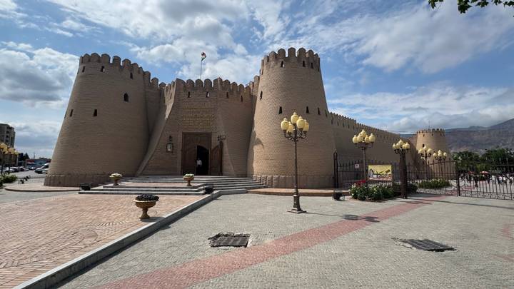Broad view of a mud-brick fortress with crenellated towers under blue skies and scattered clouds.