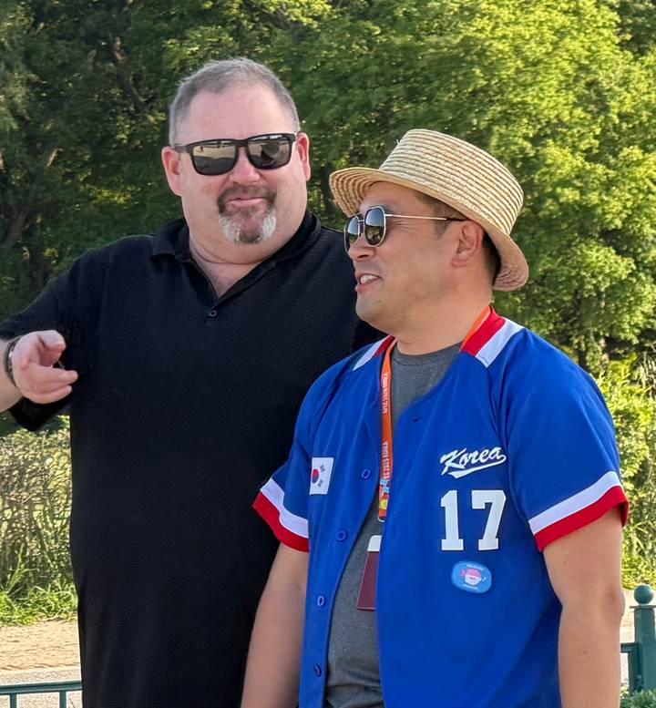 Two men smiling and talking outdoors, one wearing a straw hat and sports jersey.