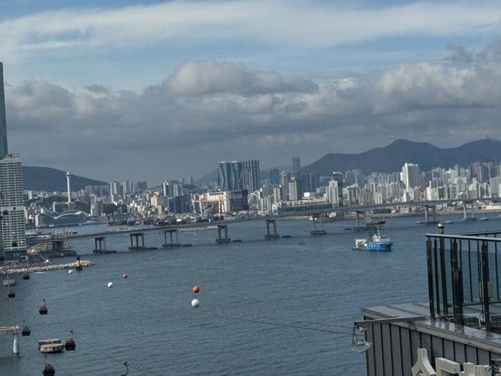 Wide harbor view of a modern city skyline with long bridge spanning a bay under scattered clouds.