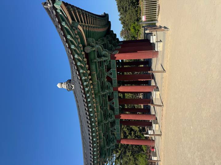 Elegant green-roofed pavilion on red pillars standing in an open sandy courtyard under clear blue sky.