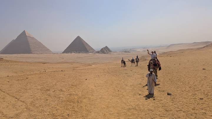 Camel riders approach the Pyramids of Giza across a vast sandy plateau under hazy skies.