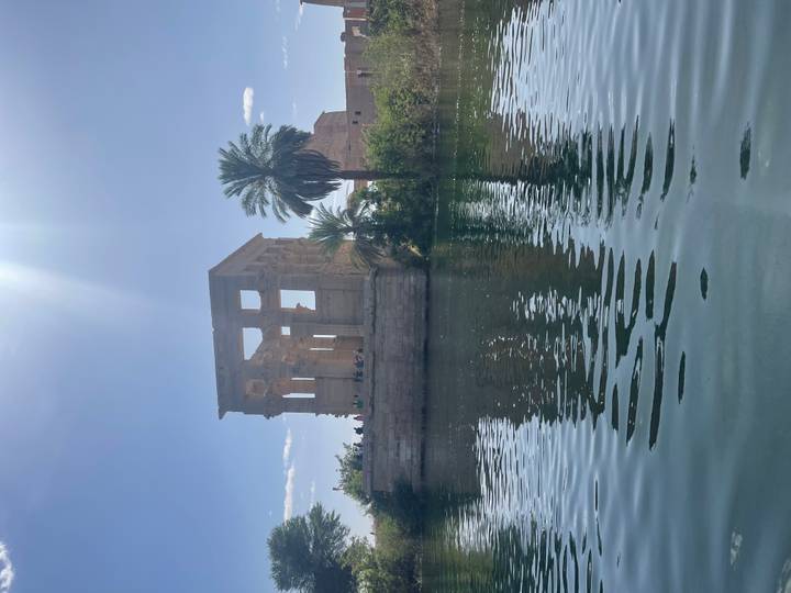 Island kiosk of Philae Temple rising from placid water under a bright midday sunbeam.