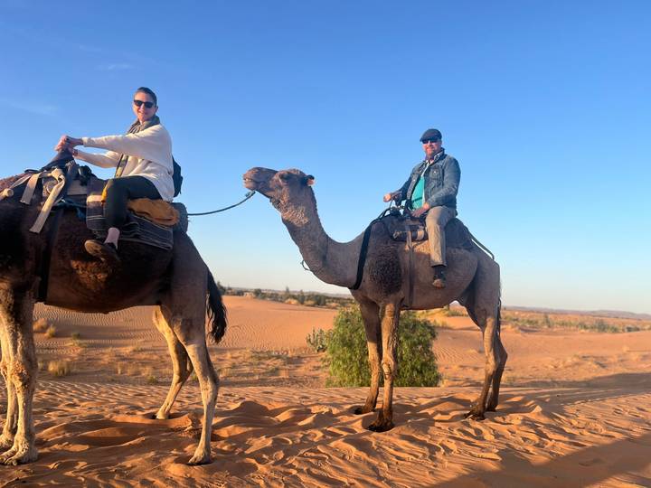 Two riders on camels against warm golden-hour light over rolling desert dunes