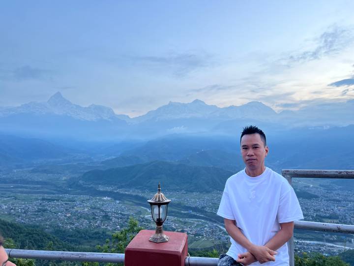 Man posing on a terrace overlooking a vast mountain valley and distant snow-covered peaks.