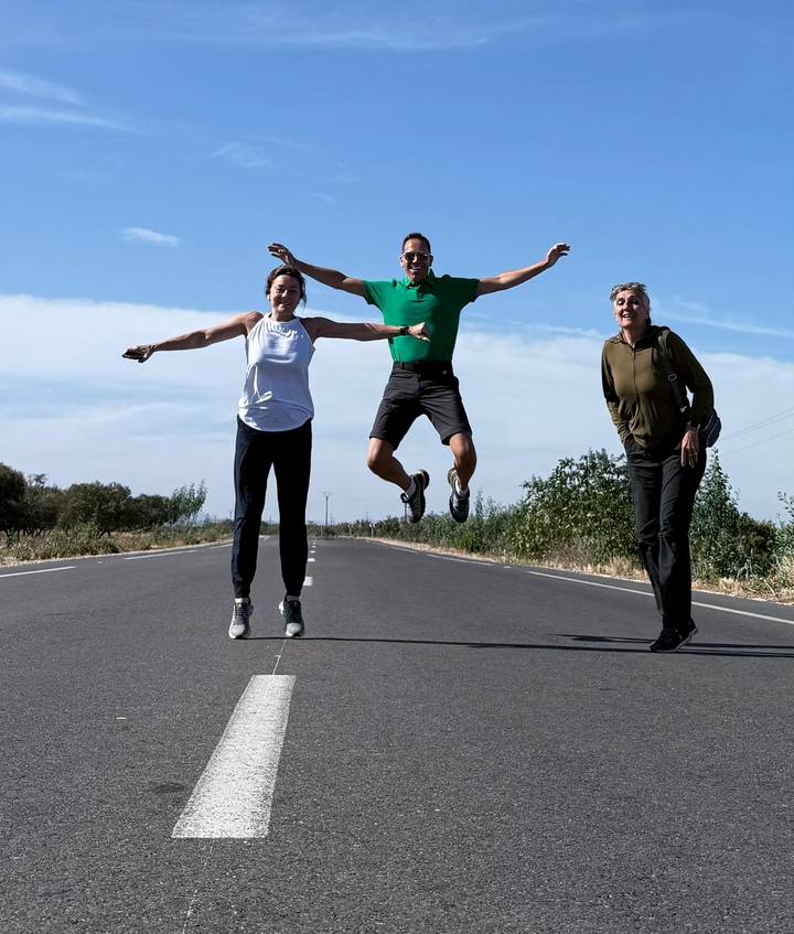 Three travellers strike playful poses, one mid-air jump, on an empty country road under a bright Moroccan sky.
