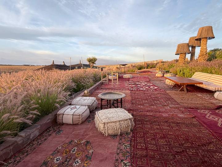 Stylish desert camp terrace decorated with patterned rugs, poufs and lanterns amid waving grass at dawn.