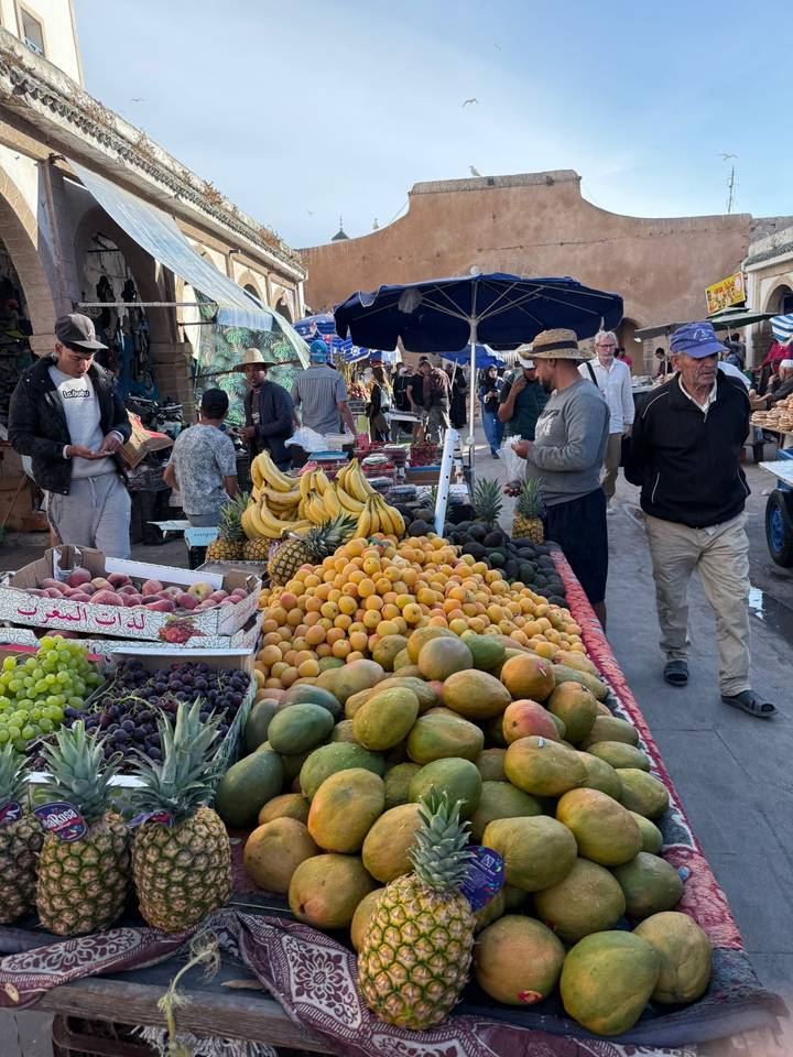 Bustling outdoor market stall piled high with colourful fruits as shoppers browse and vendors converse.