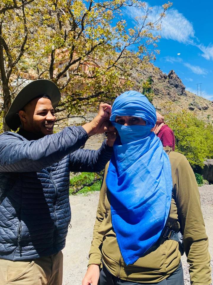 Guide helps a traveller wrap a bright blue headscarf in a mountain village setting.