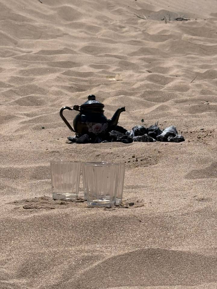 Traditional metal teapot and two glass cups rest on desert sand beside charcoal embers.