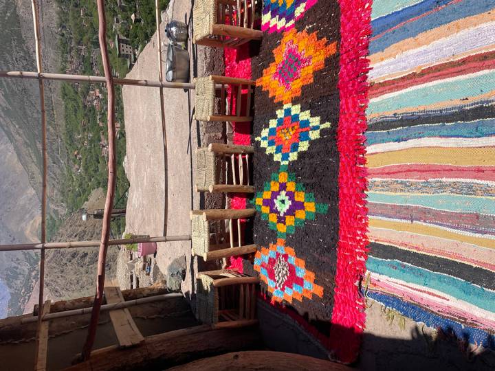 Colourful woven rugs and low stools arranged on a terrace overlooking a lush mountain valley.