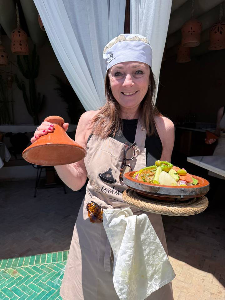 Smiling woman in apron presents a traditional Moroccan tagine dish beside its clay lid.