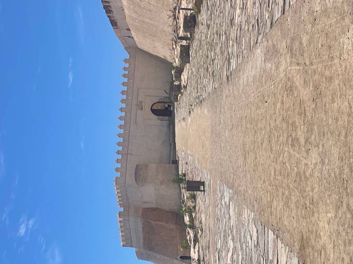 Wide sandy path leads to a crenellated fortress gate under a bright blue sky.