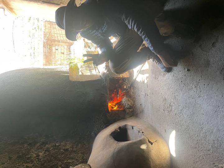 Man sitting beside a traditional clay oven with an open wood fire, flashing a peace sign in dim light.