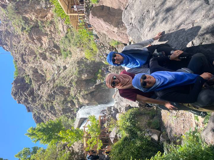 Three travellers in desert scarves pose on rocks before a cascading waterfall and rugged mountain backdrop.