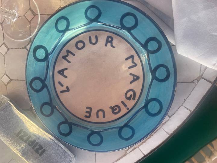 Hand-painted blue glass plate reading “L’AMOUR MAGIQUE” set on a tiled tabletop.