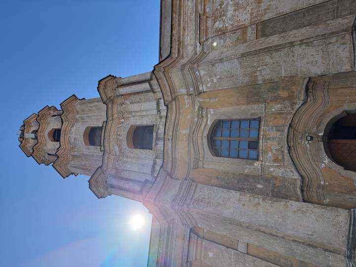 Weathered baroque church tower in Vilnius reaches into a bright blue sky with sun flare.
