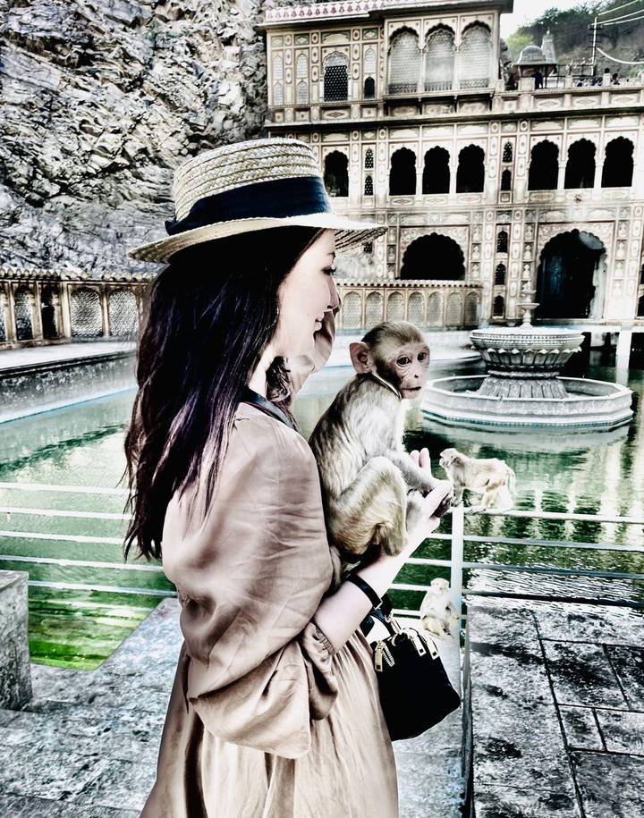 A traveller gently holds a small monkey beside an ornate pool and arched temple walls.