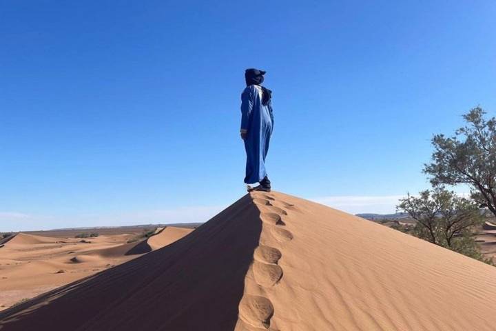 Solo traveler in blue traditional attire standing atop a sharp sand dune ridge under a clear desert sky