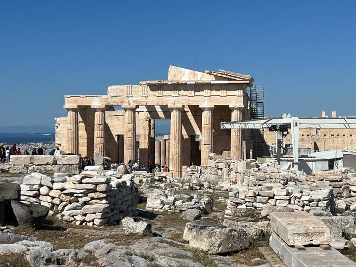 The Parthenon’s weathered columns rise above scattered ancient stones with tourists exploring the Acropolis plateau.