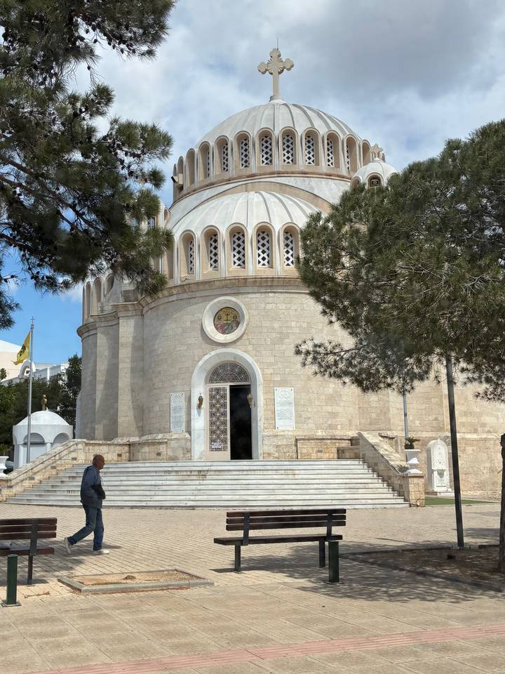 A circular stone church with ornate windows and a small dome is framed by pine branches; a man sits on the steps.
