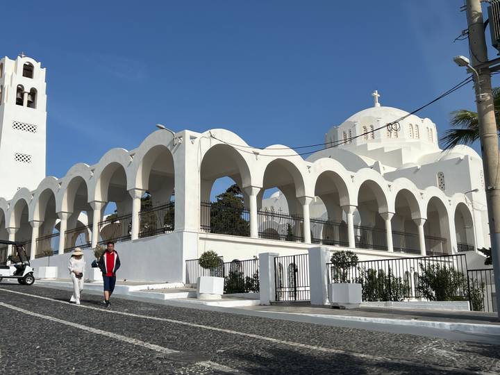 Two people walk past a large white domed church with multiple arches on a sunny island street.