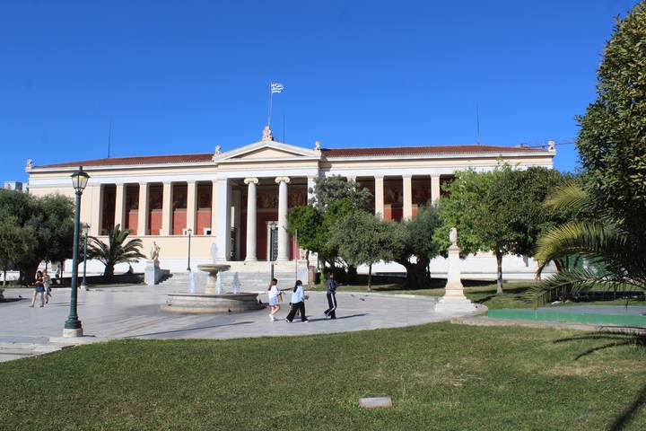 A neoclassical university building with red columns fronts a fountain and green lawn where people stroll.