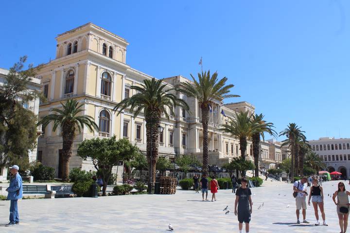 Elegant neoclassical town hall with palm trees and people enjoying a sunny square on a Greek island.