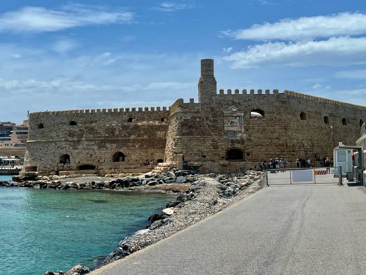 A robust Venetian stone fortress juts into turquoise harbor waters with a breakwater walkway leading up to it.