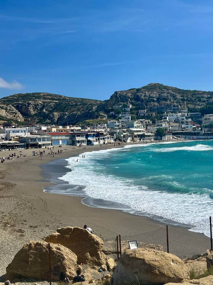 A coastal town backed by rocky hills curves around a sandy beach where turquoise waves crash and sunbathers relax.
