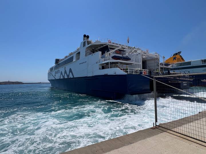 A large high-speed passenger ferry prepares to depart, churning blue water at the stern beside a pier fence.