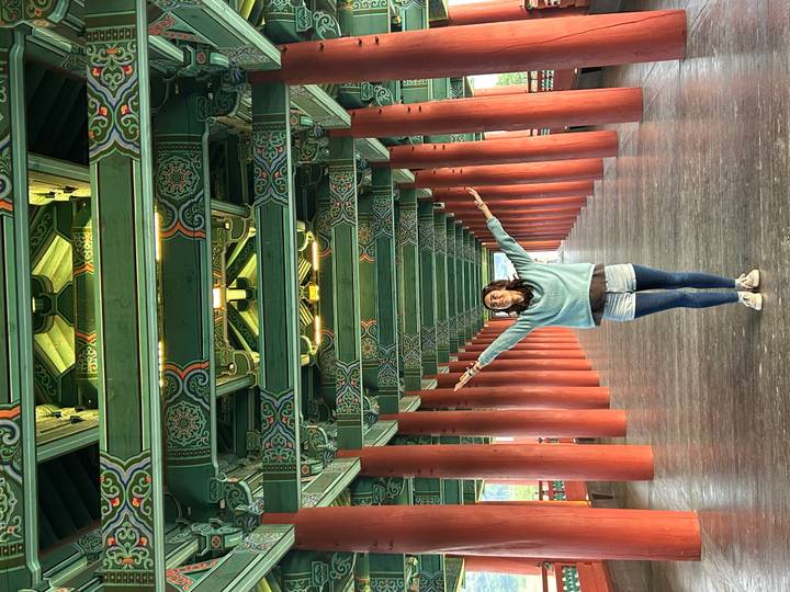Young traveller standing arms wide in a vast green-painted wooden hall lined with red columns.