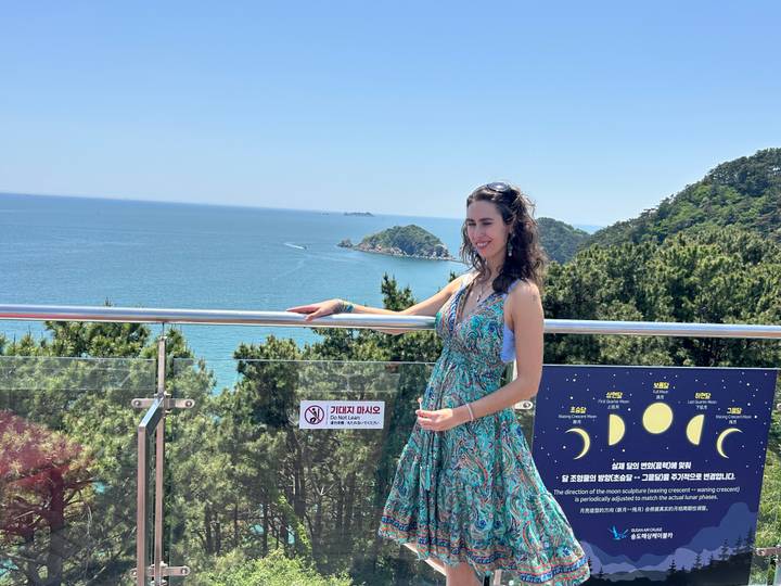 Woman in summer dress enjoying a coastal panorama from a glass balcony above the blue sea.