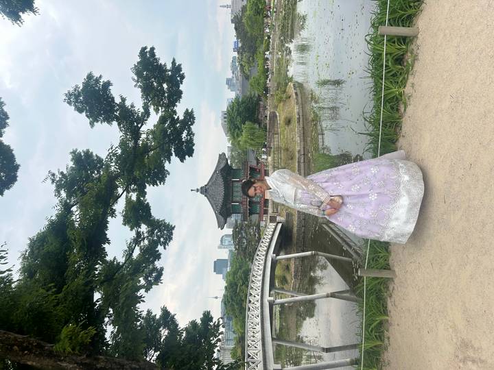 Woman wearing pastel hanbok posing near a traditional pavilion and pond in central Seoul.